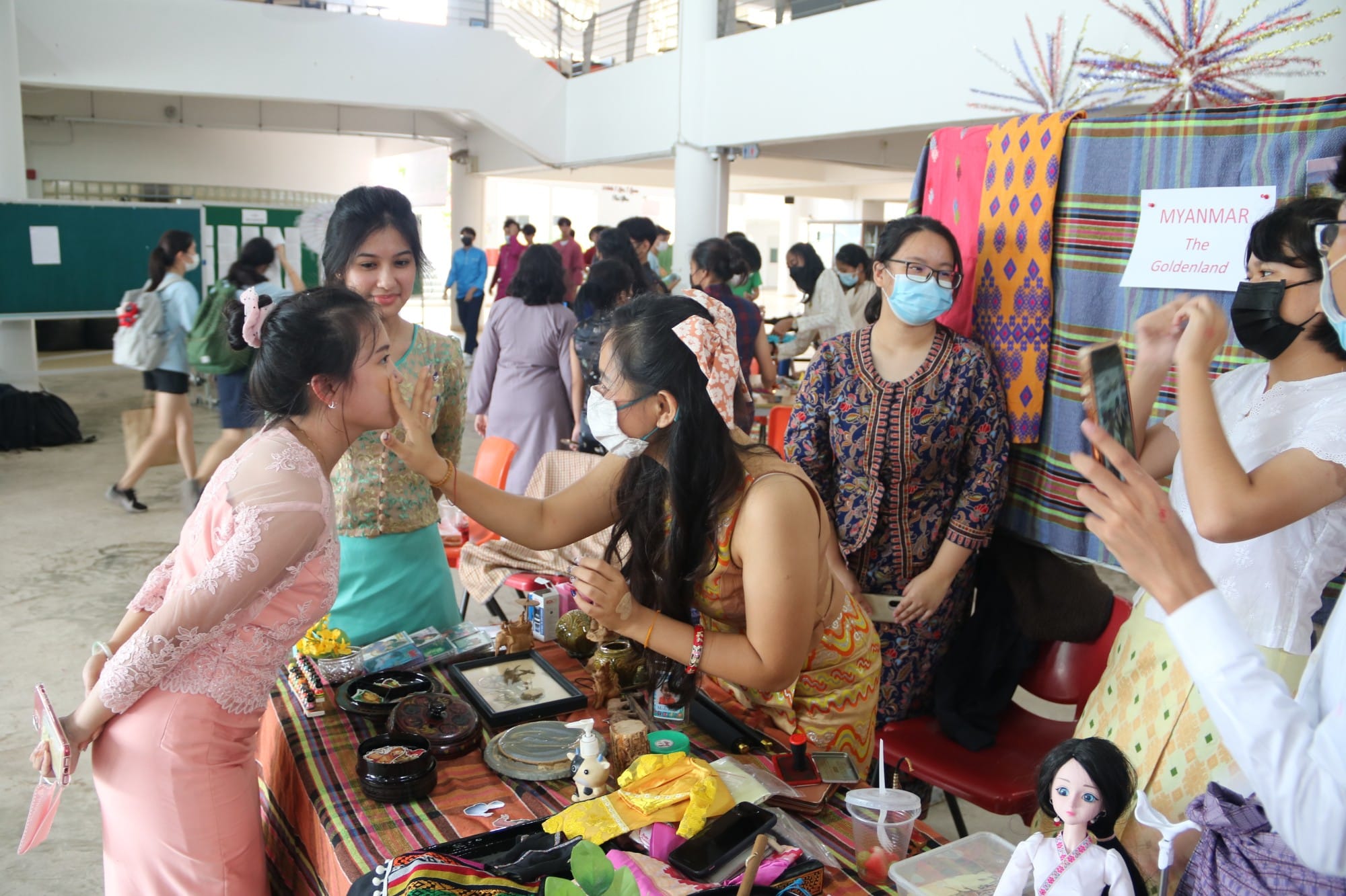 People in traditional clothing gathered around a Myanmar cultural booth displaying crafts, fabrics, and dolls at an indoor fair.